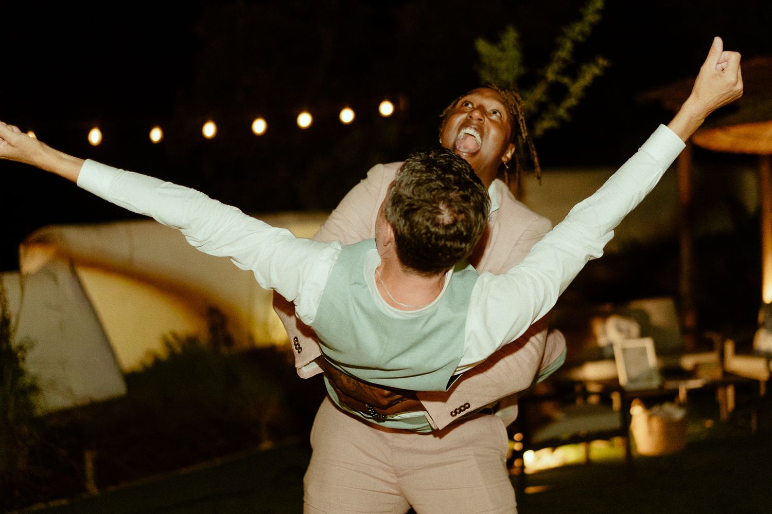 Couple celebrating outdoors at night, woman with arms outstretched on man's back, string lights glowing behind them.
