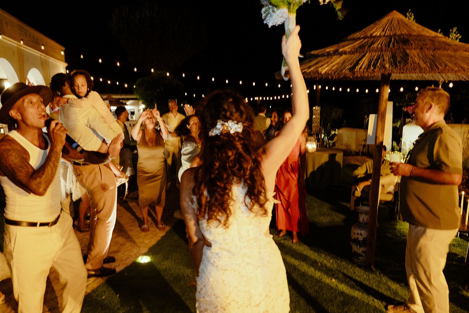 Bride tosses bouquet at outdoor wedding reception at night with string lights and guests celebrating.