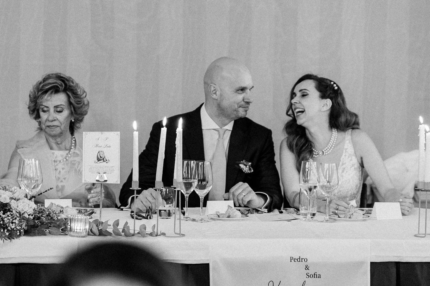 Black and white wedding photo of smiling bride and groom seated at reception table with candles and champagne glasses.
