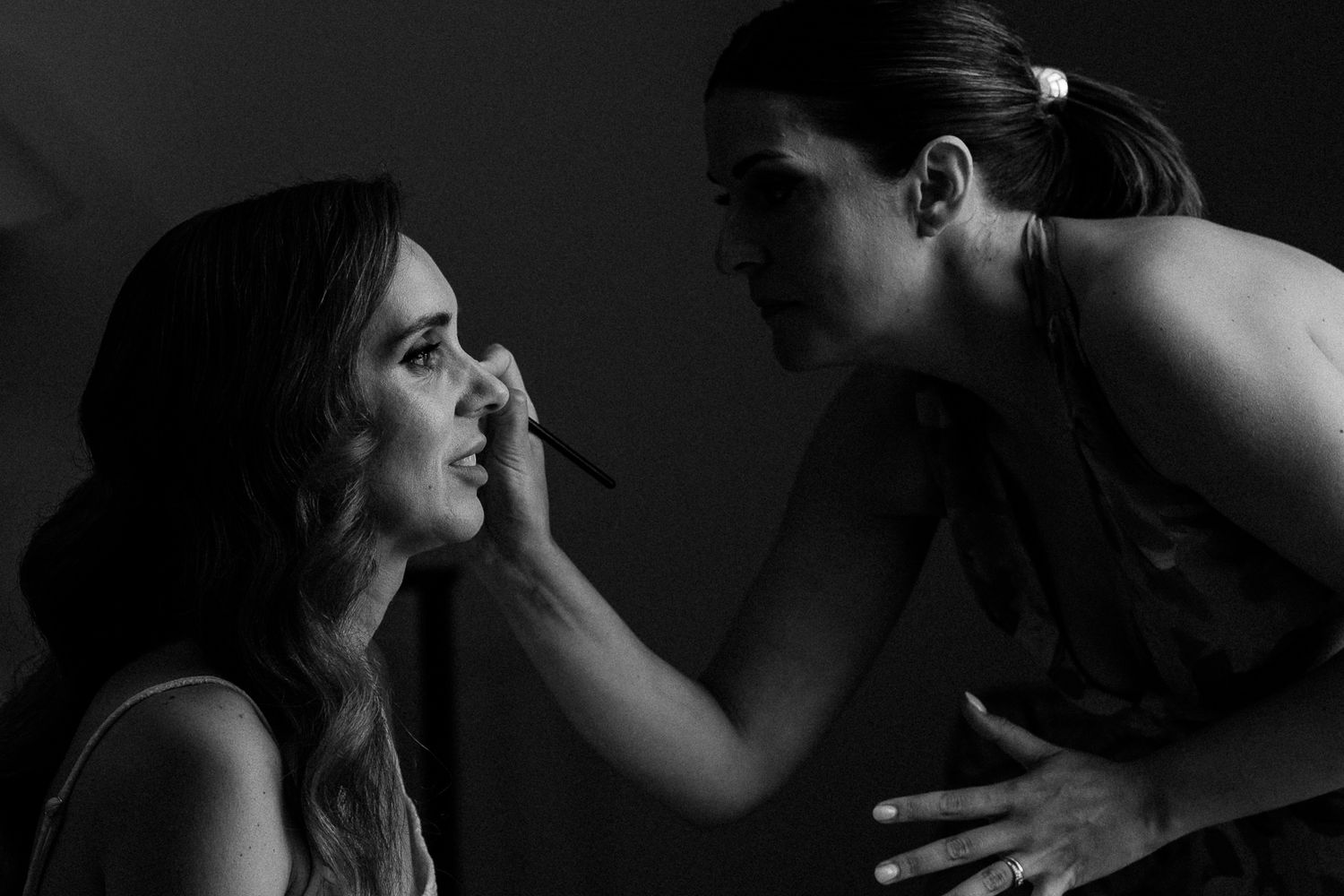 Black and white photo of a makeup artist applying makeup to a woman in a dimly lit studio.