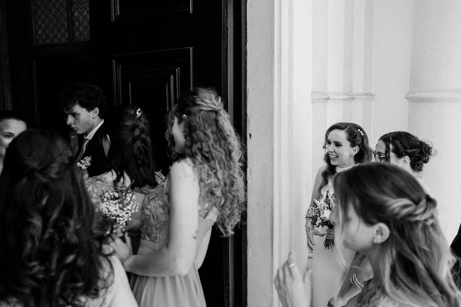 Black and white photo of bridesmaids gathering near a doorway, smiling and chatting before a wedding ceremony.
