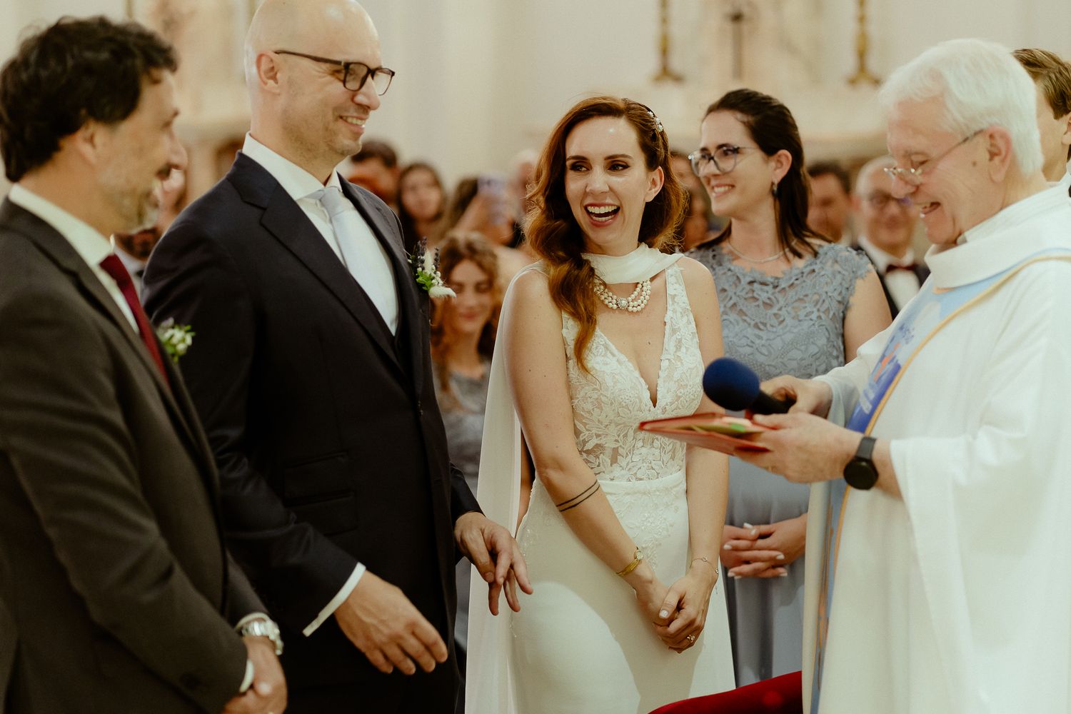 Bride and groom smiling during wedding ceremony with priest holding book in elegant church setting.