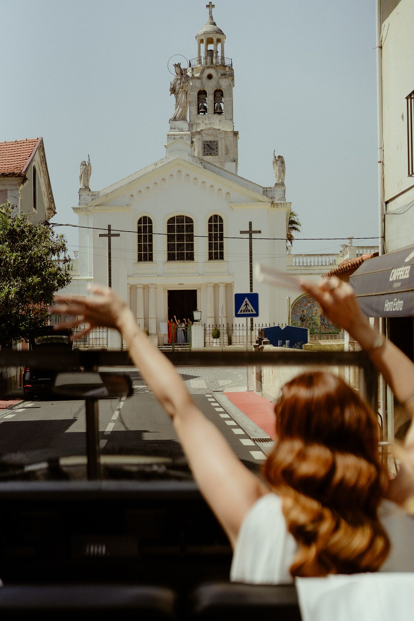 Woman with raised arms facing a white historic church with bell tower on a sunny European street.