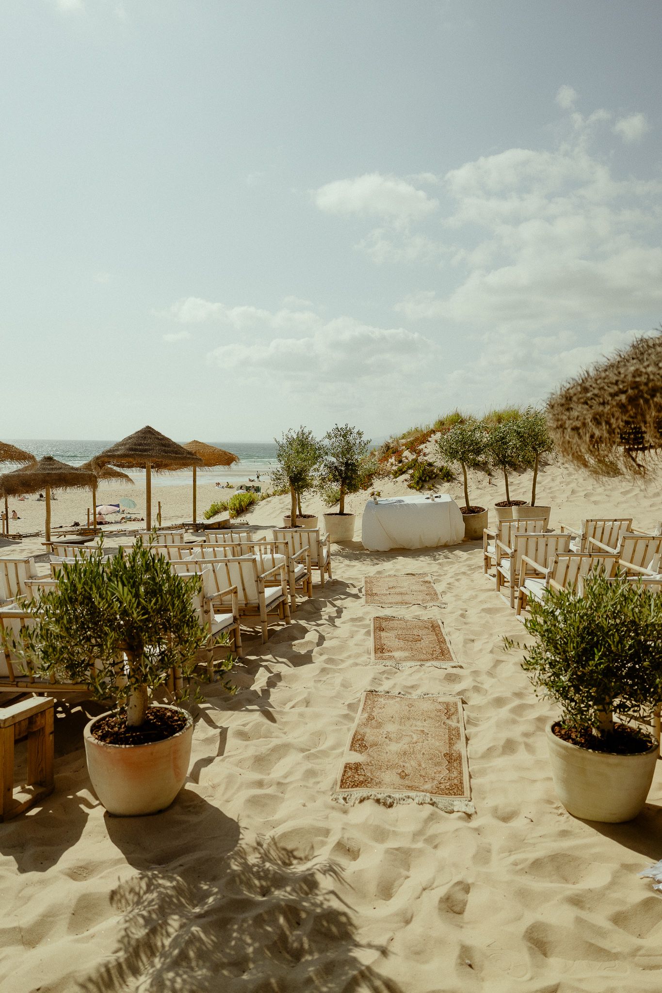 Sandy beach setup with white chairs, potted olive trees, vintage rugs, and straw umbrellas creating an elegant outdoor event space.