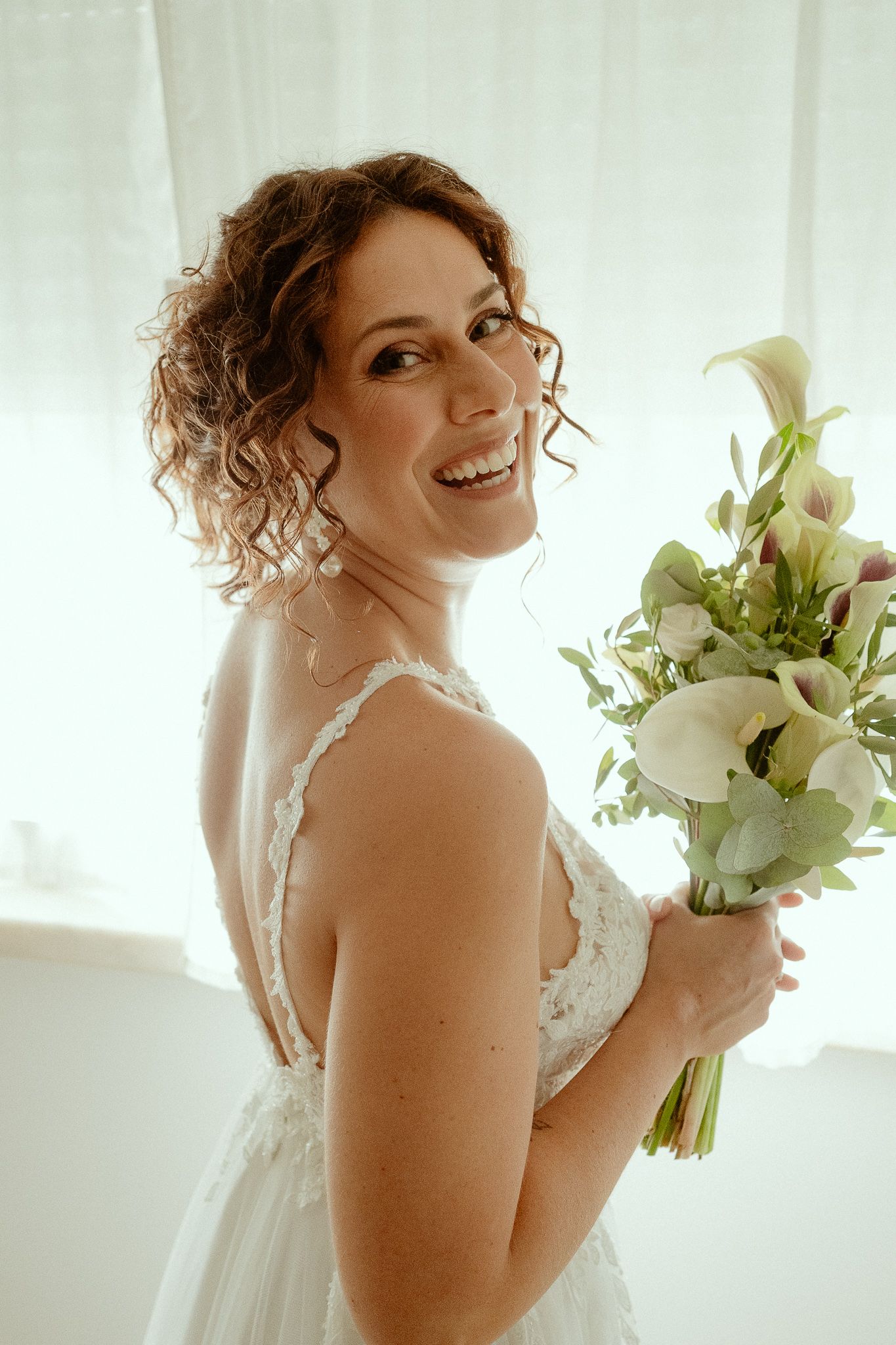 Smiling bride in white lace dress holding calla lily bouquet, posing elegantly in bright natural light.