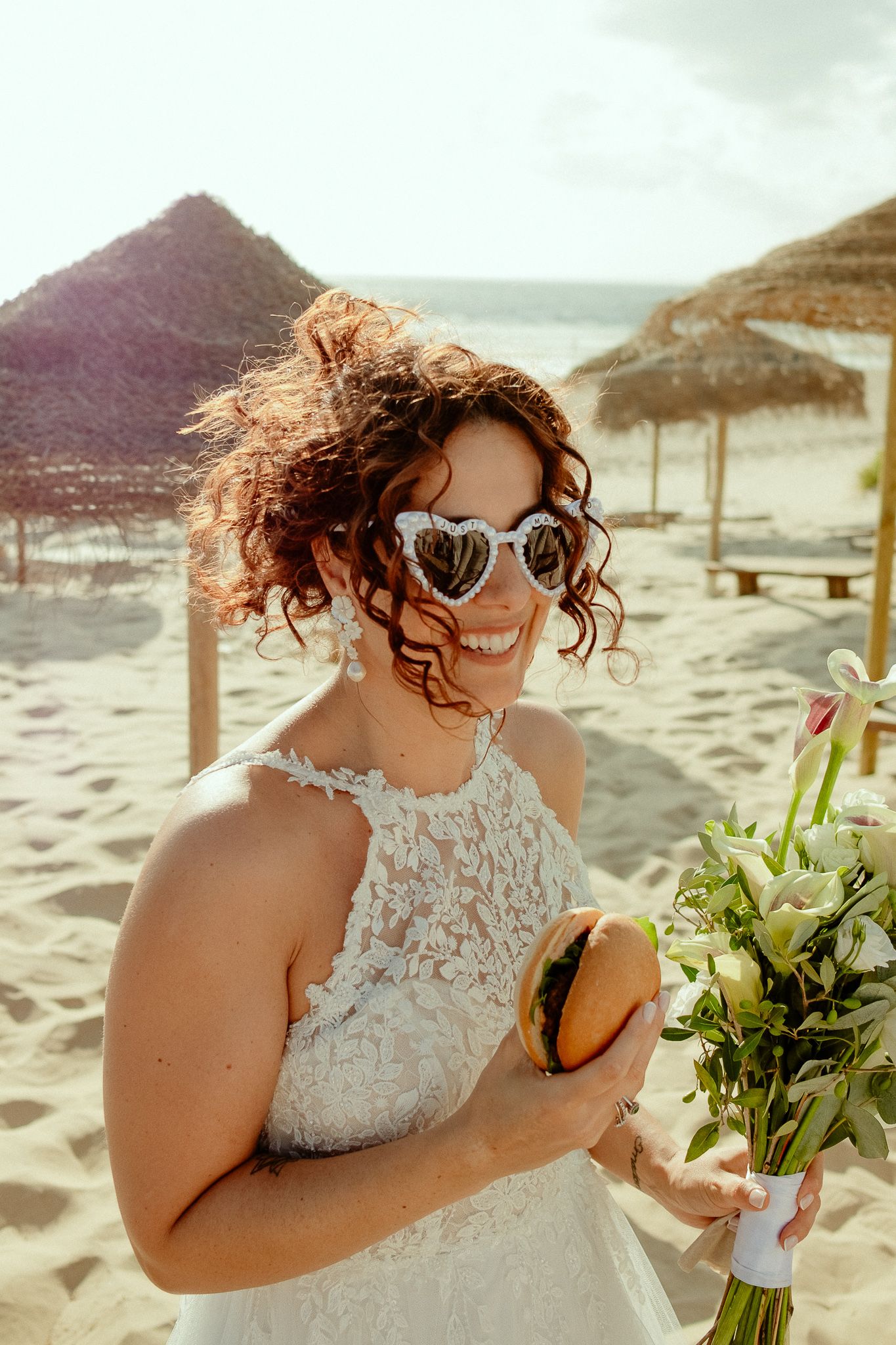 Smiling bride in white lace dress and heart-shaped sunglasses holds bouquet and burger on sandy beach.