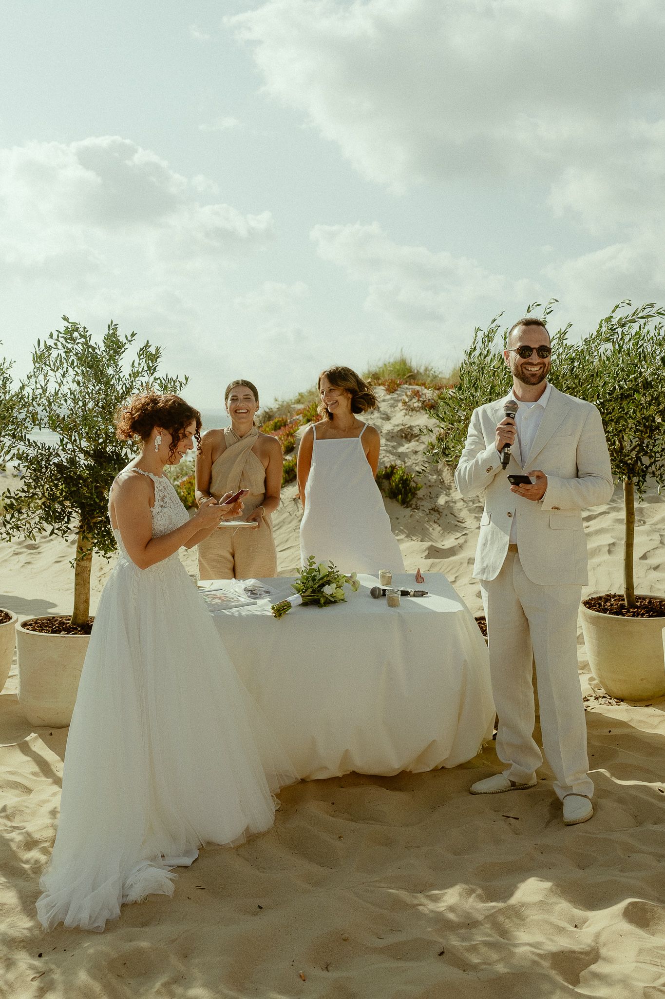 Couple exchanges vows at beach wedding ceremony with guests and olive trees on sandy dunes under cloudy sky.