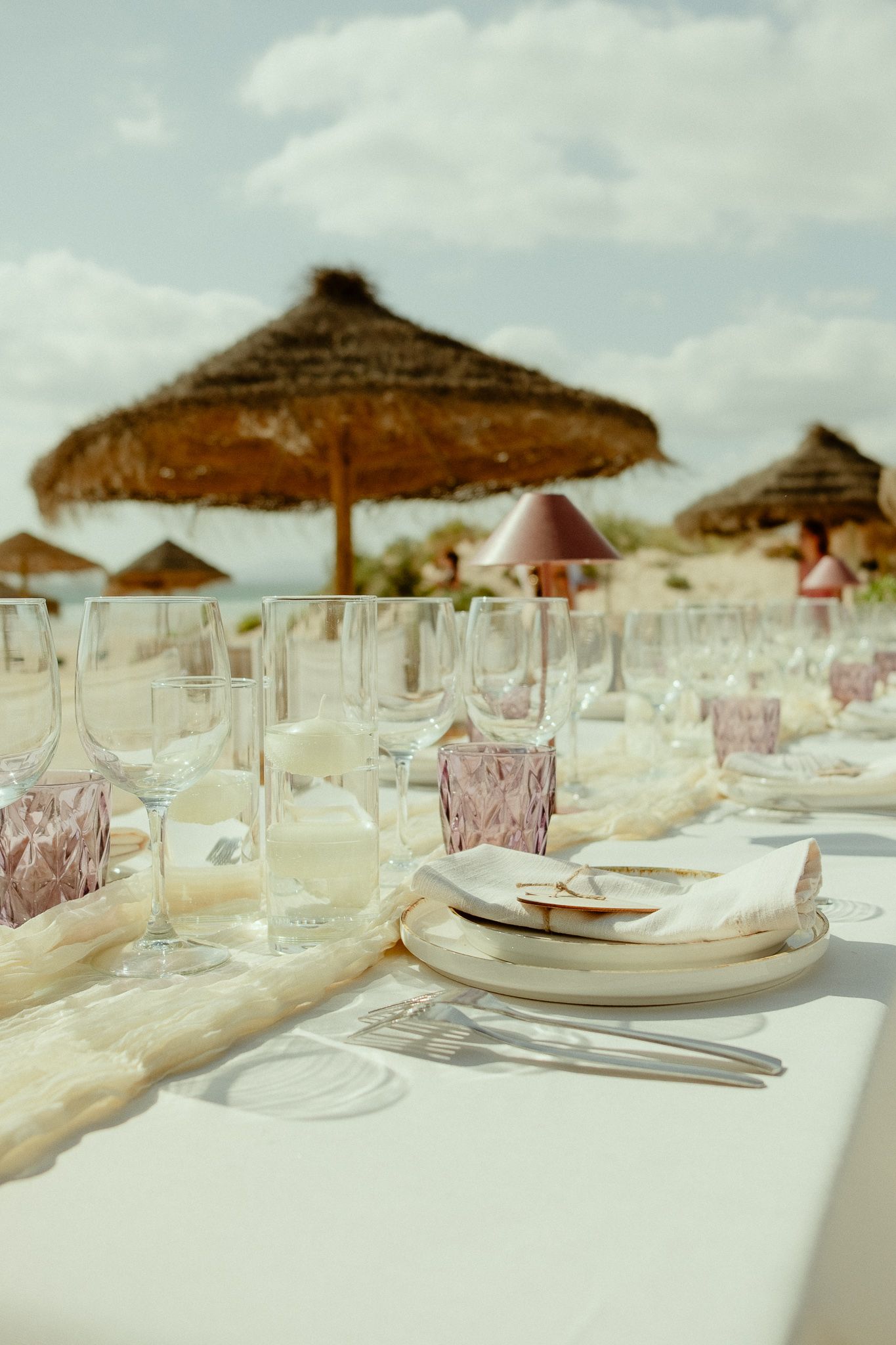 Elegant beach dining table set with crystal glasses, white linens, and thatched umbrellas under a cloudy sky.