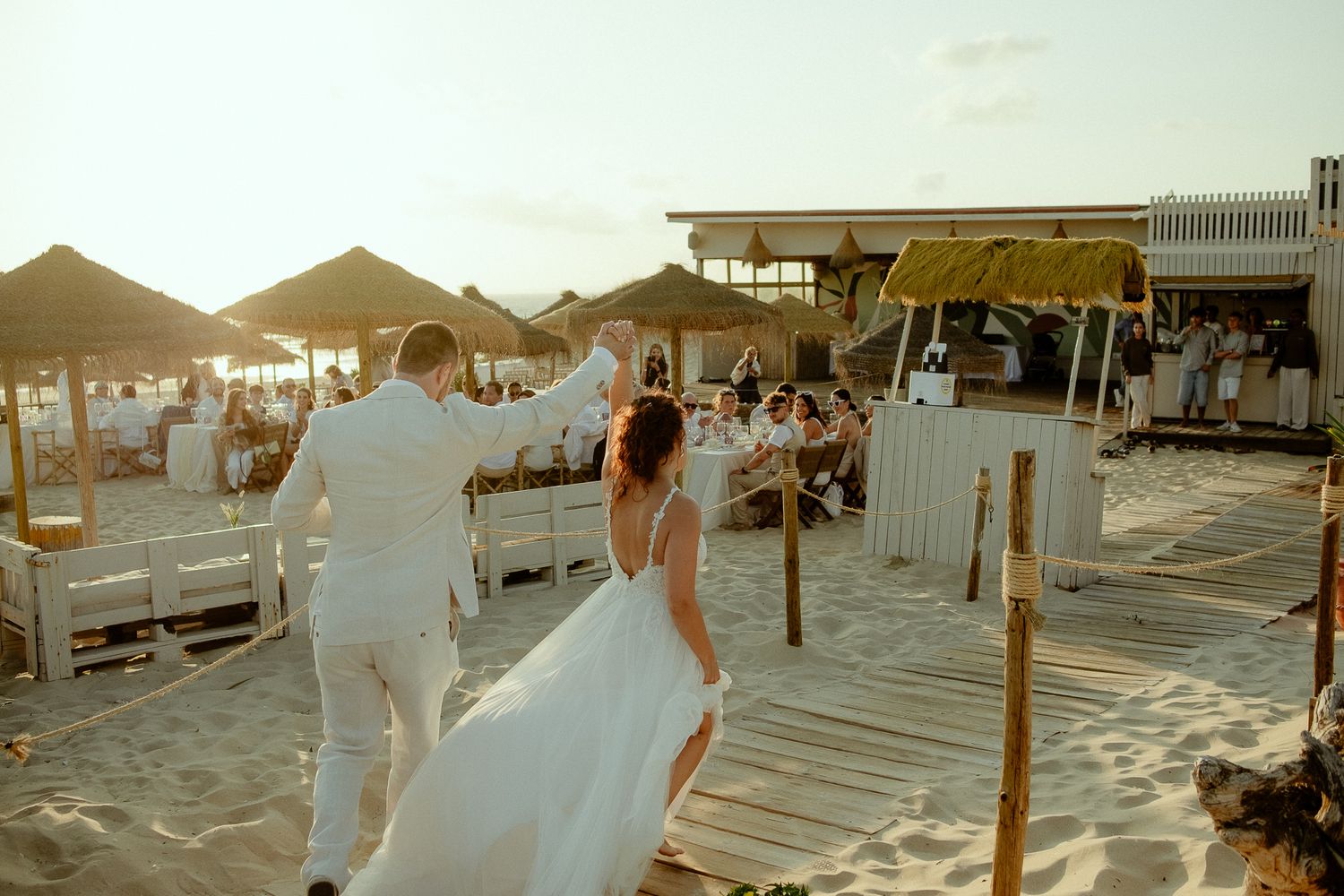 Bride and groom dancing on sandy beach venue at sunset with white decor and thatched umbrellas.
