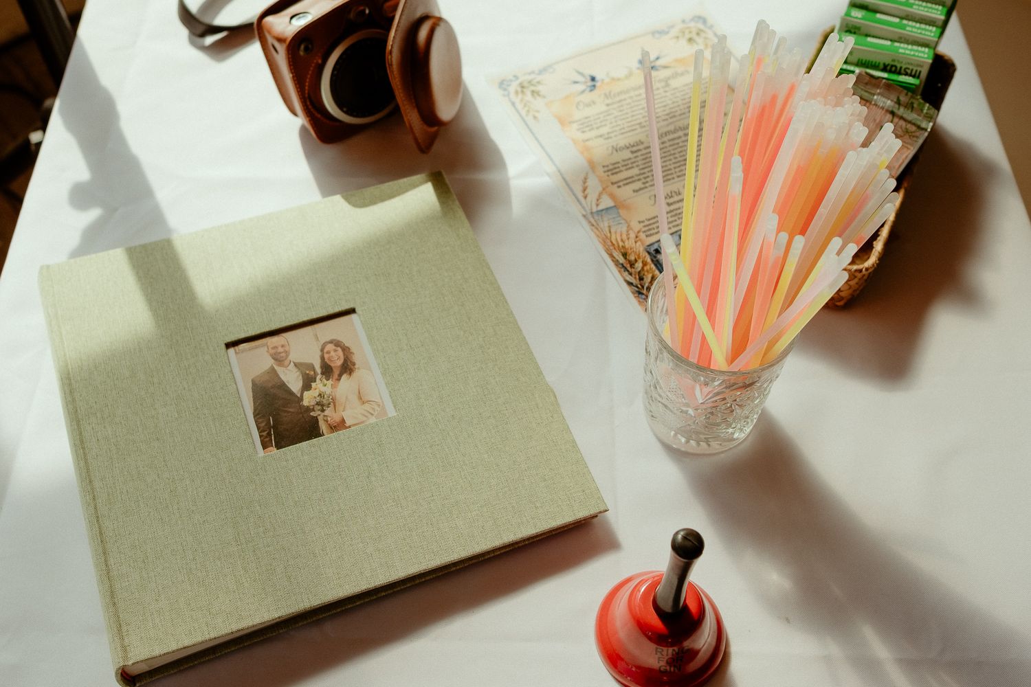 A wedding photo album beside a vintage camera and a glass vase with colorful straws on a white table.