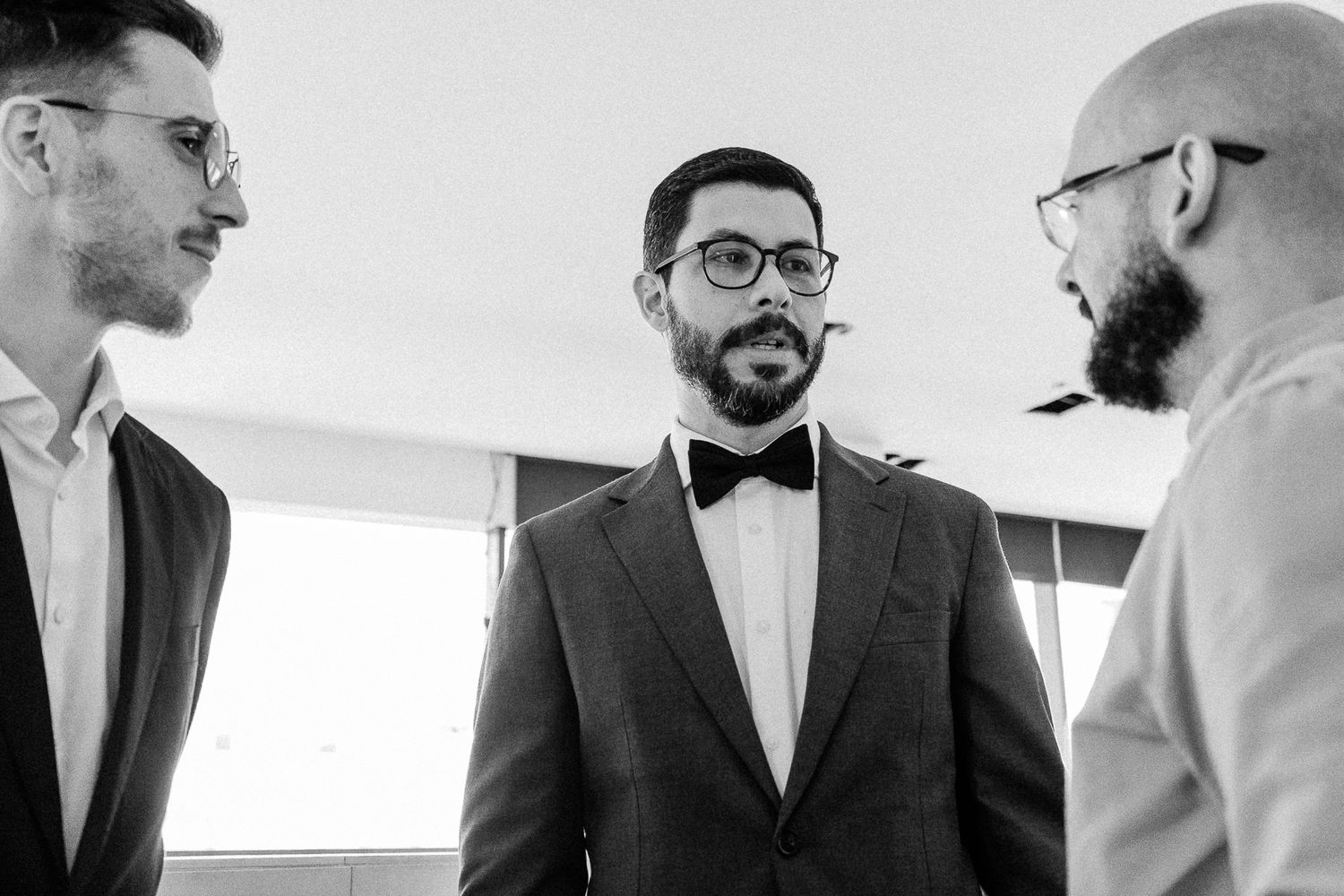 Black and white photo of three men in formal attire, groom in bow tie chatting with groomsmen before wedding.