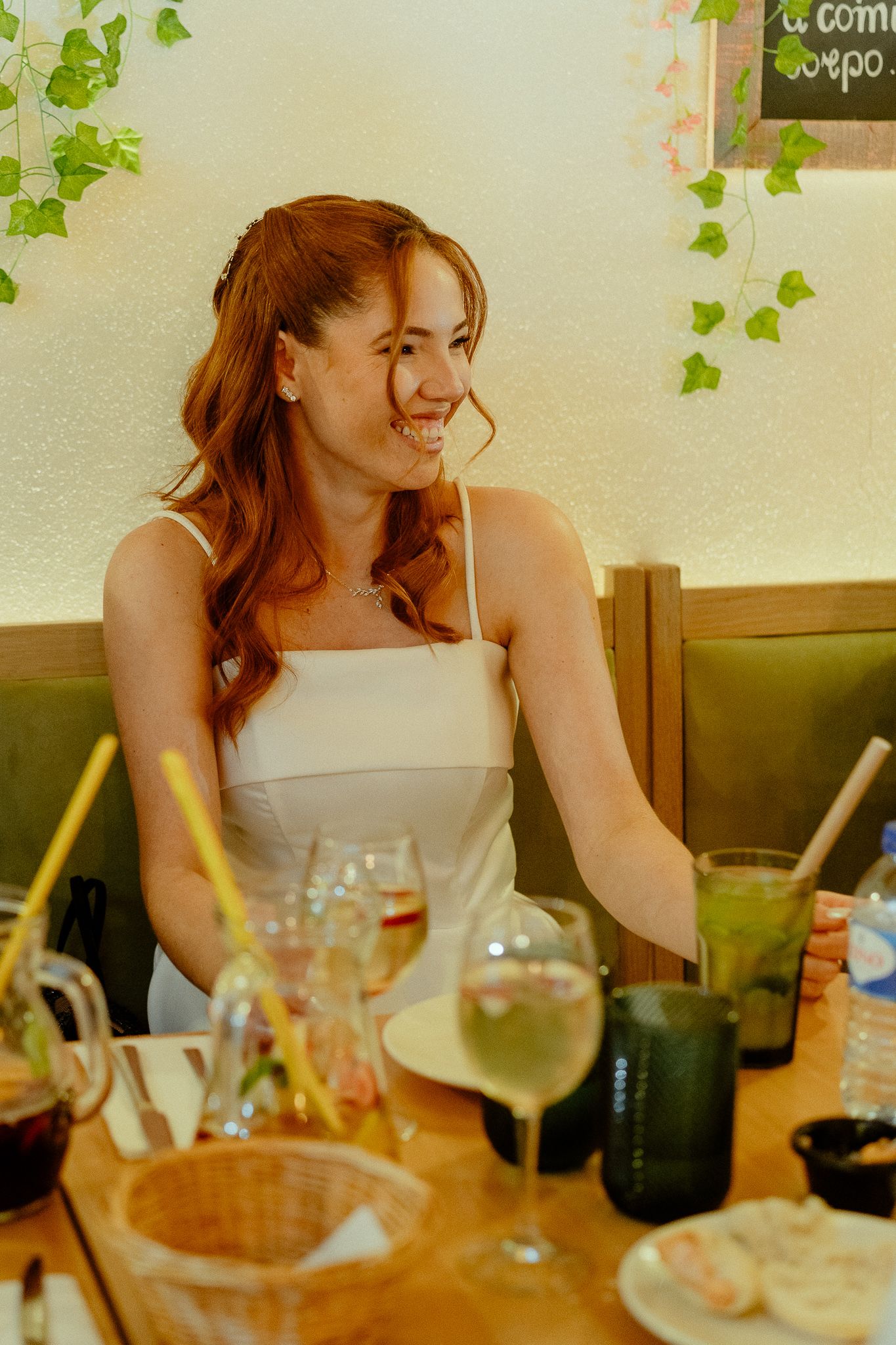 Smiling redhead woman in white dress sitting at restaurant table with drinks and greenery decor.