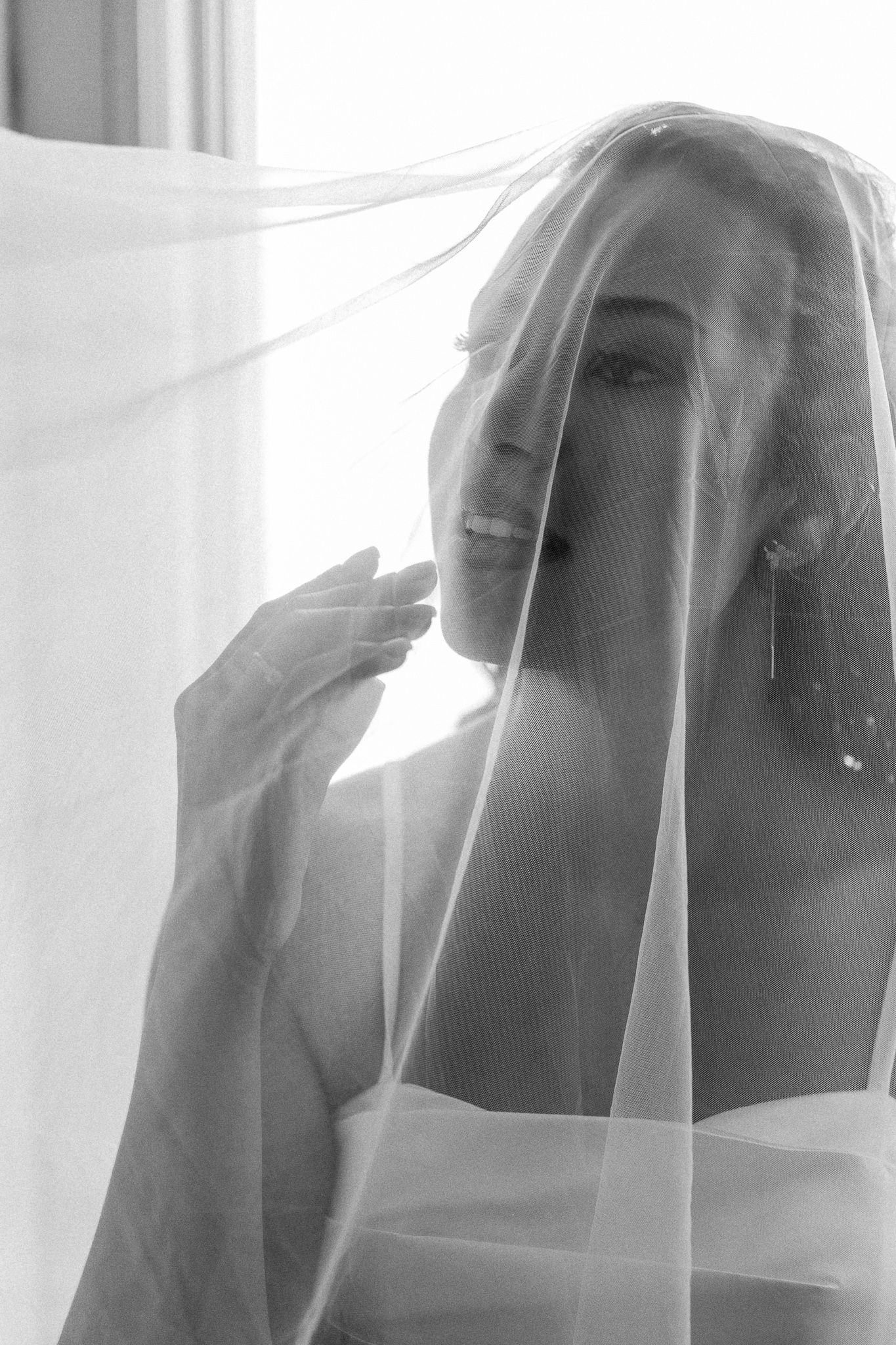 Bride draped in a sheer veil poses in soft light in black and white photo.