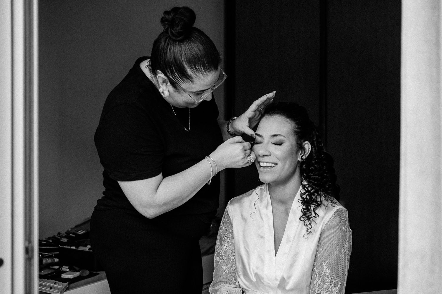Black and white photo of a makeup artist applying eyebrow makeup to a smiling bride in a white robe.