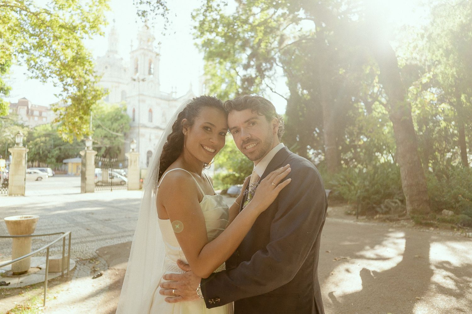 A smiling bride and groom embrace outdoors on their wedding day with sunlit trees and a church in the background.