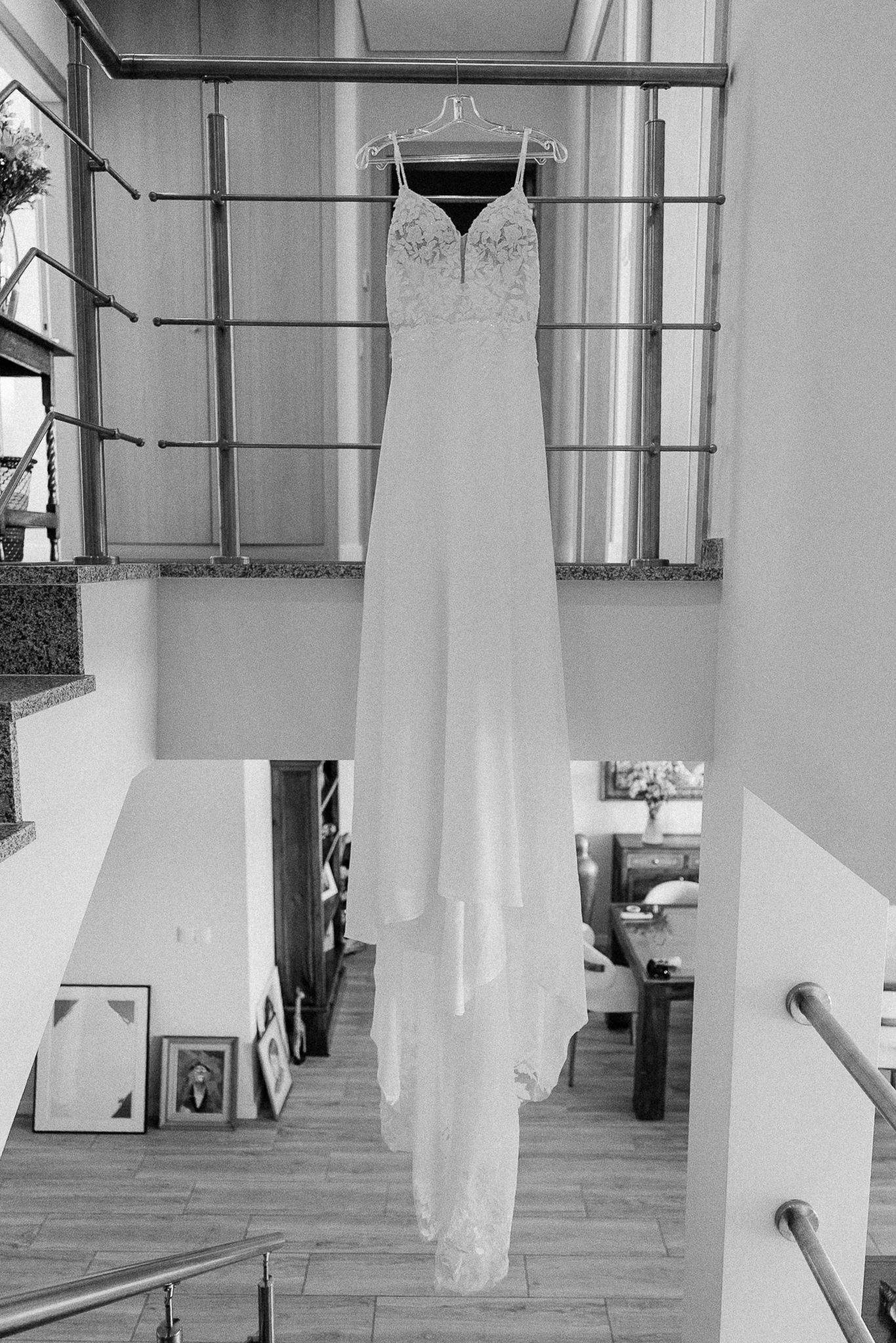 Black and white photo of an elegant wedding dress hanging from a balcony railing in a modern two-story home.