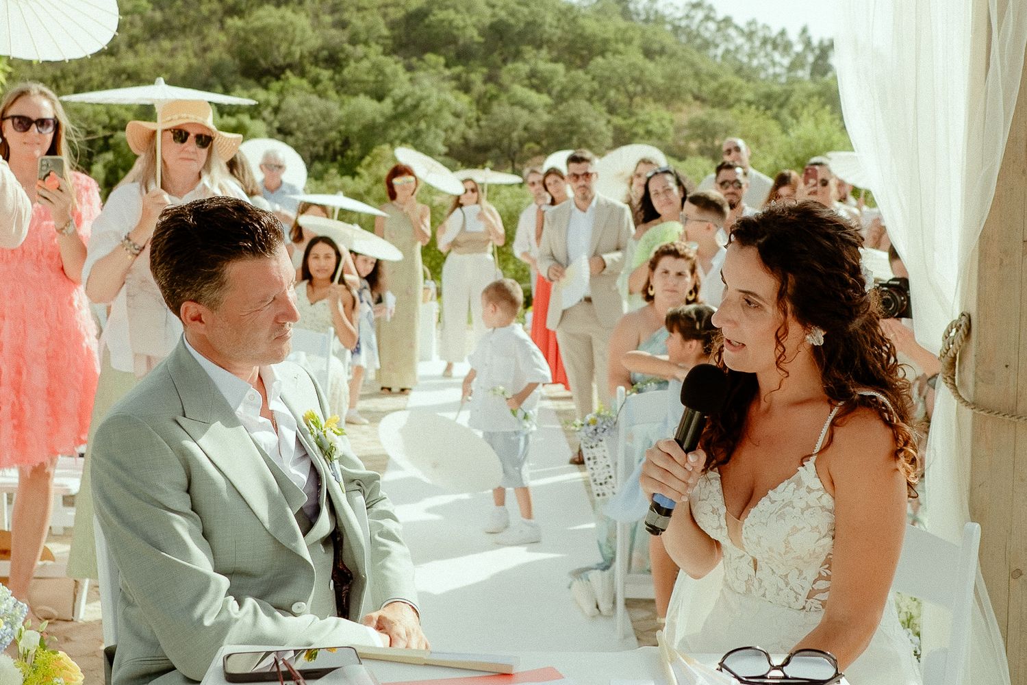Bride reads vows to groom at outdoor wedding ceremony with guests watching in background.