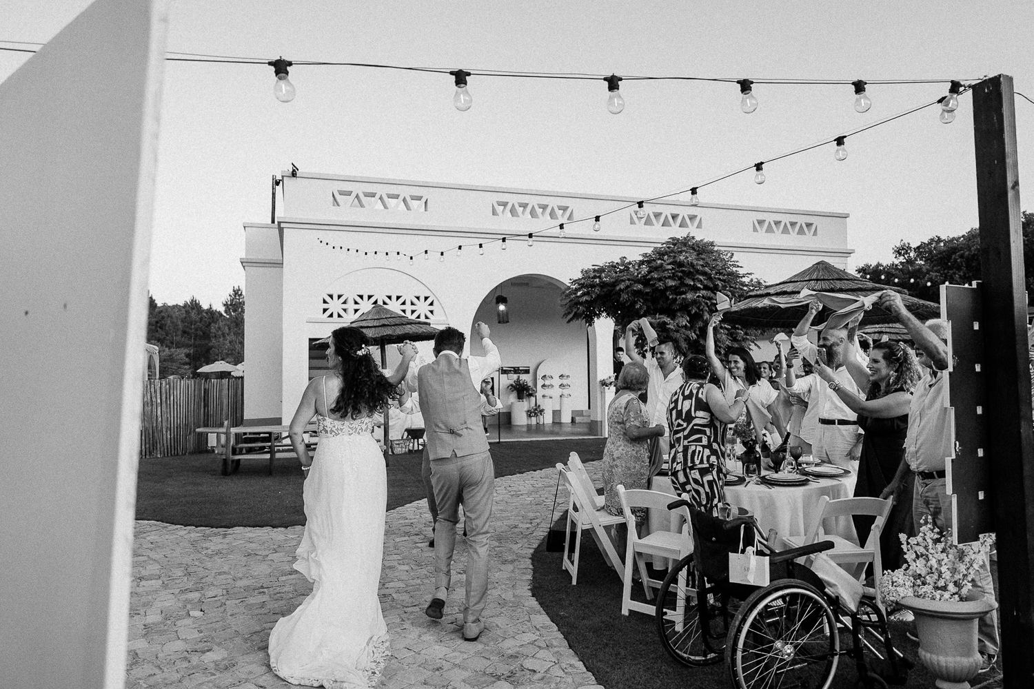 Black and white photo of a bride and groom entering an outdoor wedding reception with string lights and guests.