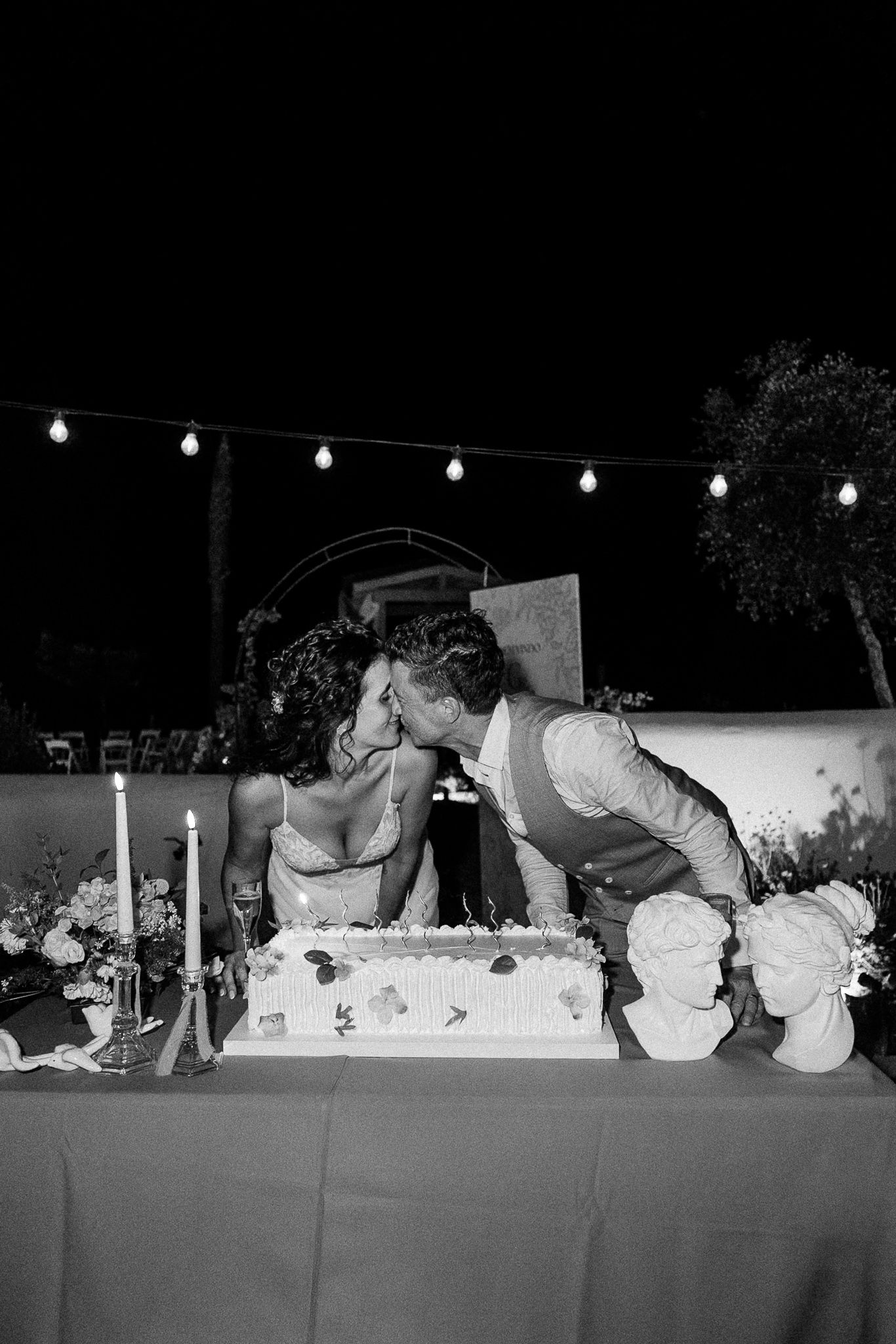 Black and white photo of a couple kissing over their wedding cake at an outdoor night reception with string lights.
