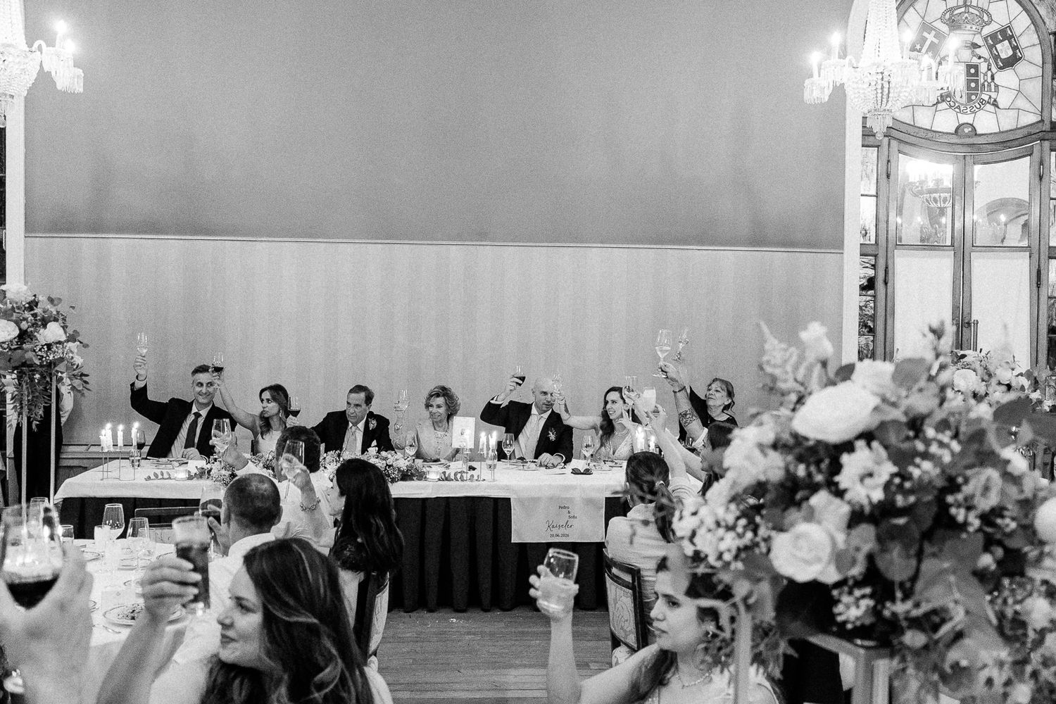 Black and white wedding reception toast with guests raising glasses toward the head table adorned with floral arrangements.