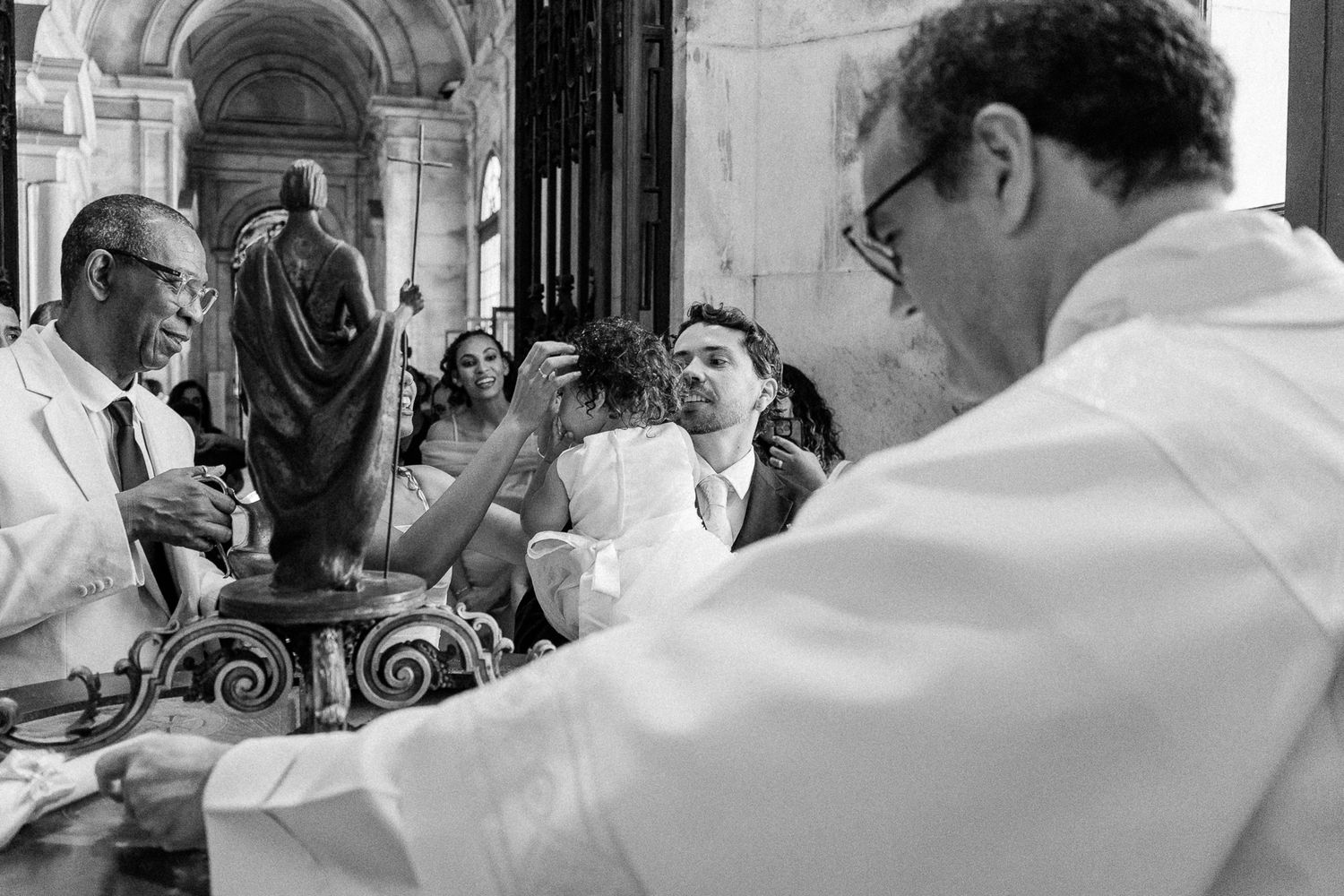 Black and white photo of a Catholic baptism ceremony with priest and attendees gathered around a font in a church.