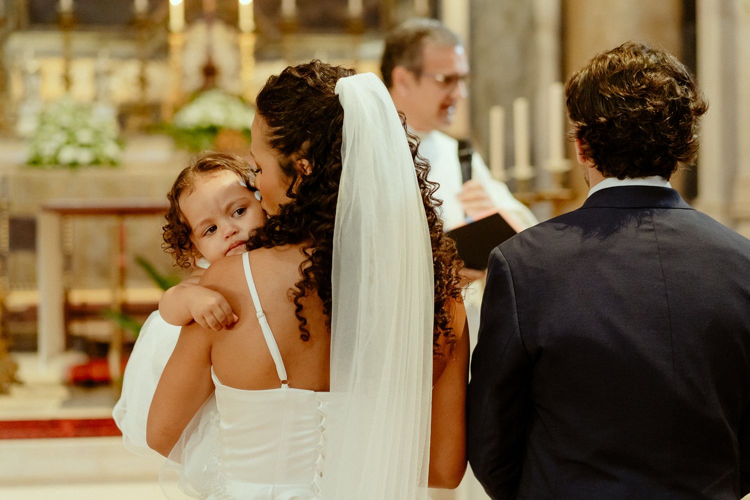 Bride in white gown holds child at church altar during wedding ceremony with groom and officiant nearby.