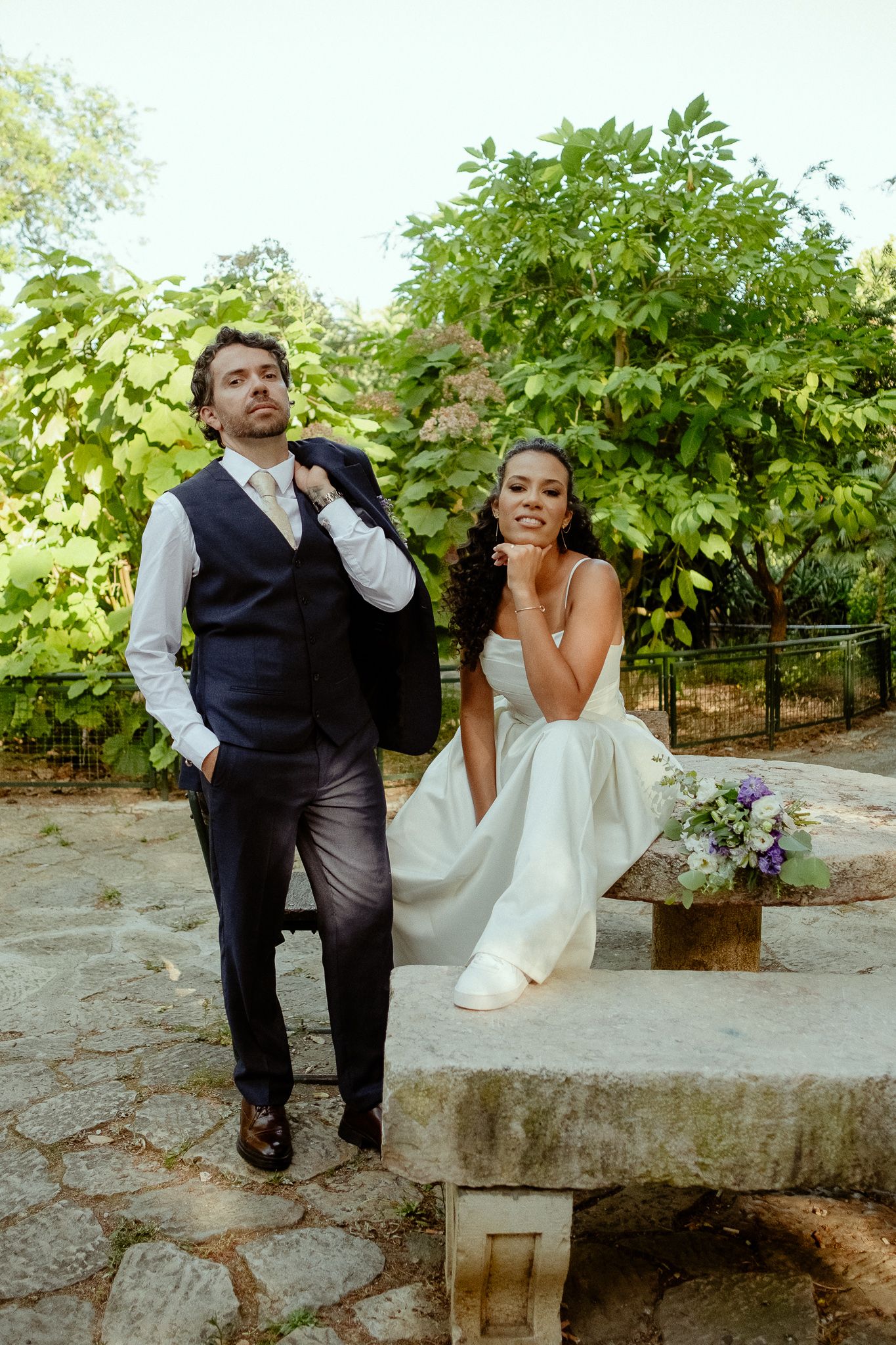 A bride and groom pose outdoors in a garden, she sits on a stone bench with a bouquet as he adjusts his suit jacket.