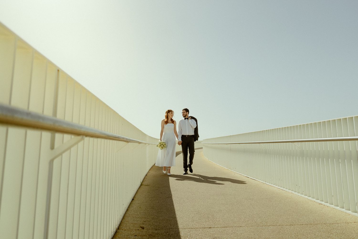 Couple walking hand in hand on a bright modern bridge pathway, woman in white dress, man in black suit.