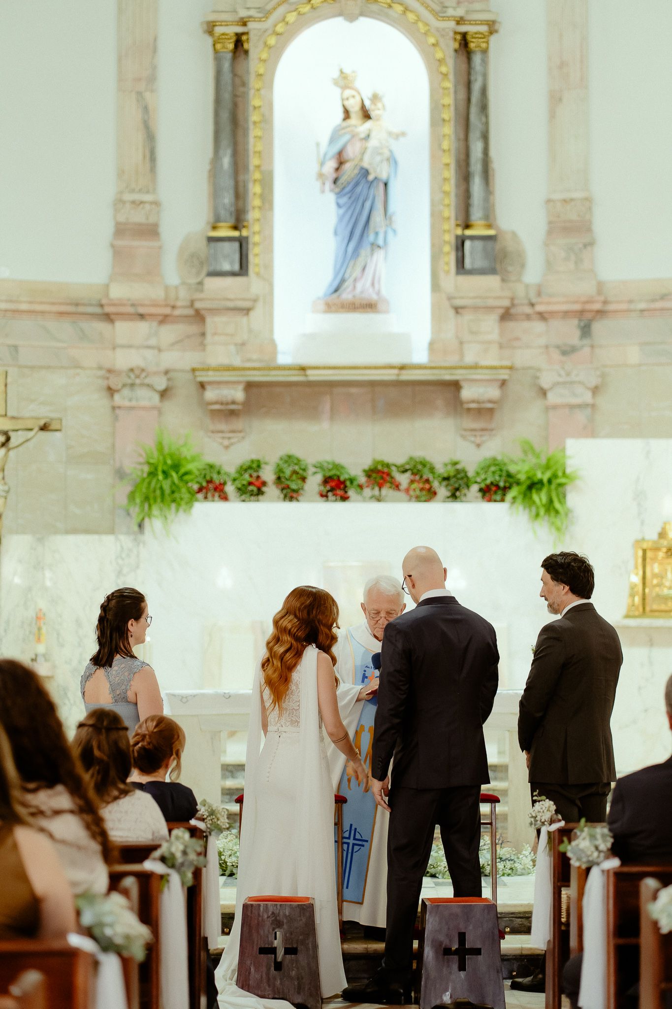 Bride and groom exchanging vows at a Catholic church altar with a Virgin Mary statue and floral decorations.