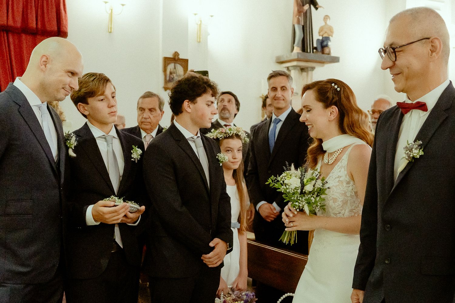 Bride and groom exchange vows at church wedding ceremony surrounded by groomsmen and flower girl holding bouquet.