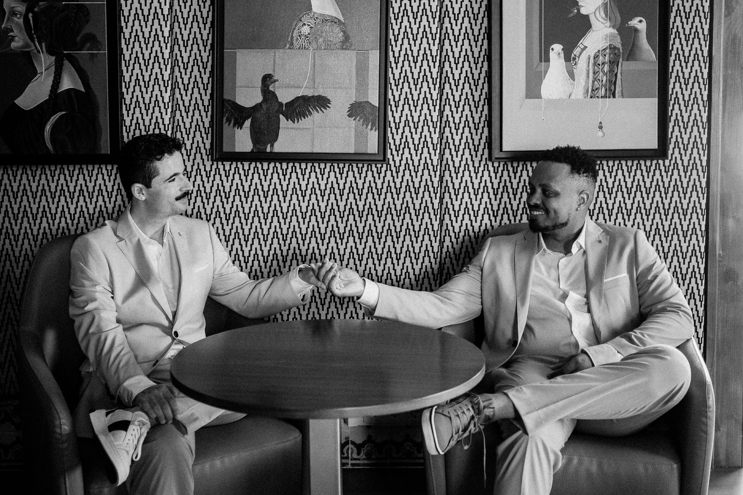 Two men in light suits shake hands and smile at a round table in a stylish black and white photo.