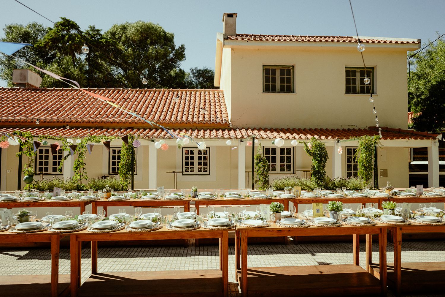 Elegant outdoor wedding reception with long wooden tables set for dining in front of a charming villa with red tile roof.