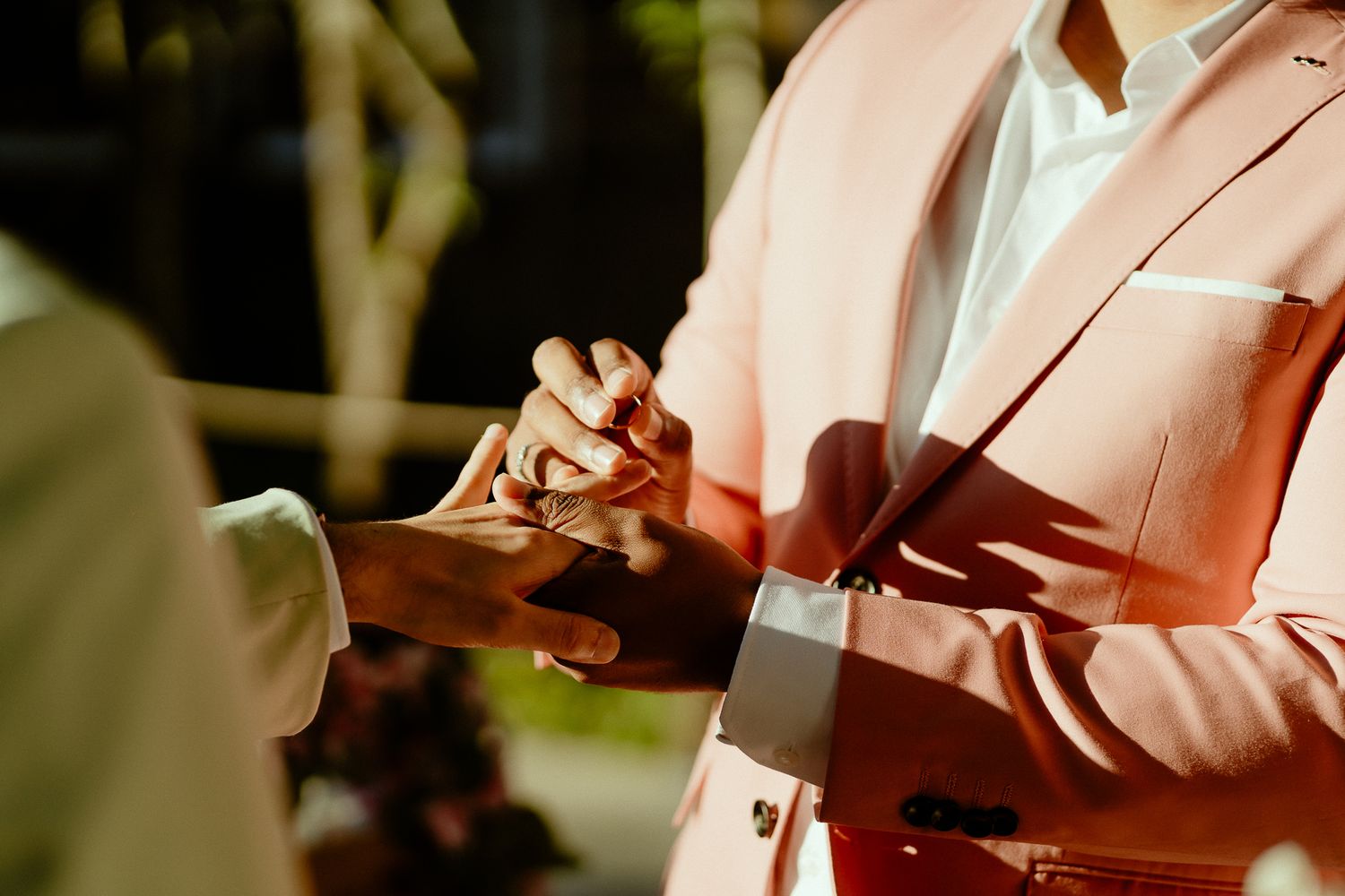 Groom in pink suit placing wedding ring on partner's finger during outdoor ceremony.