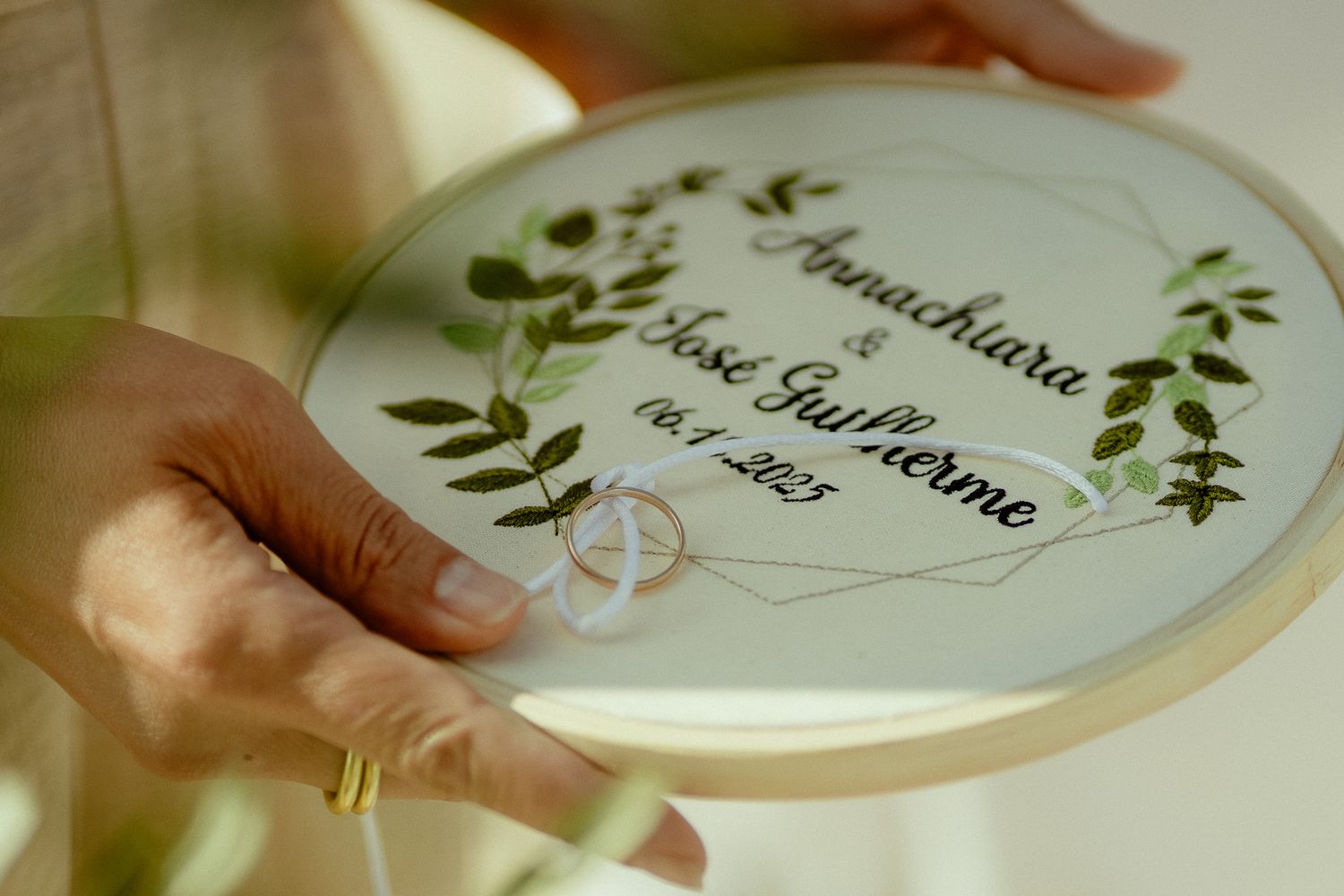 Hands holding an embroidery hoop wedding ring bearer with floral design and names Annachiara and Jose Guilherme.