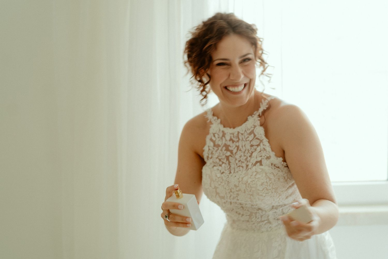 Smiling bride in a white lace halter wedding dress laughing joyfully while getting ready indoors.