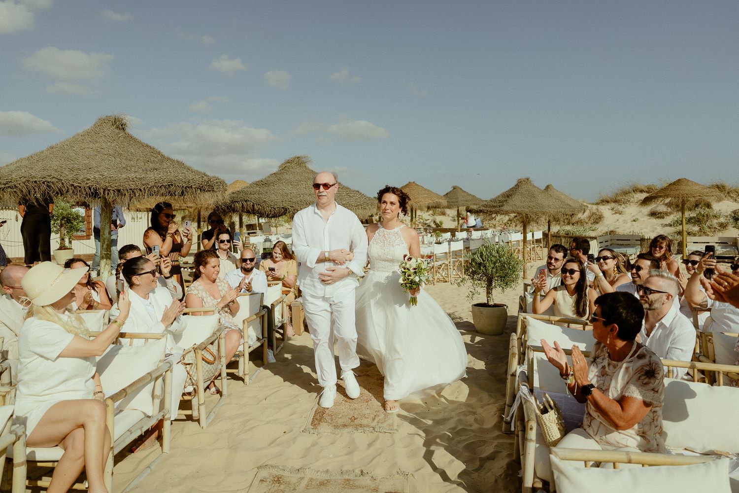 Bride and groom walk down sandy beach aisle surrounded by guests under thatched umbrellas at outdoor wedding ceremony.