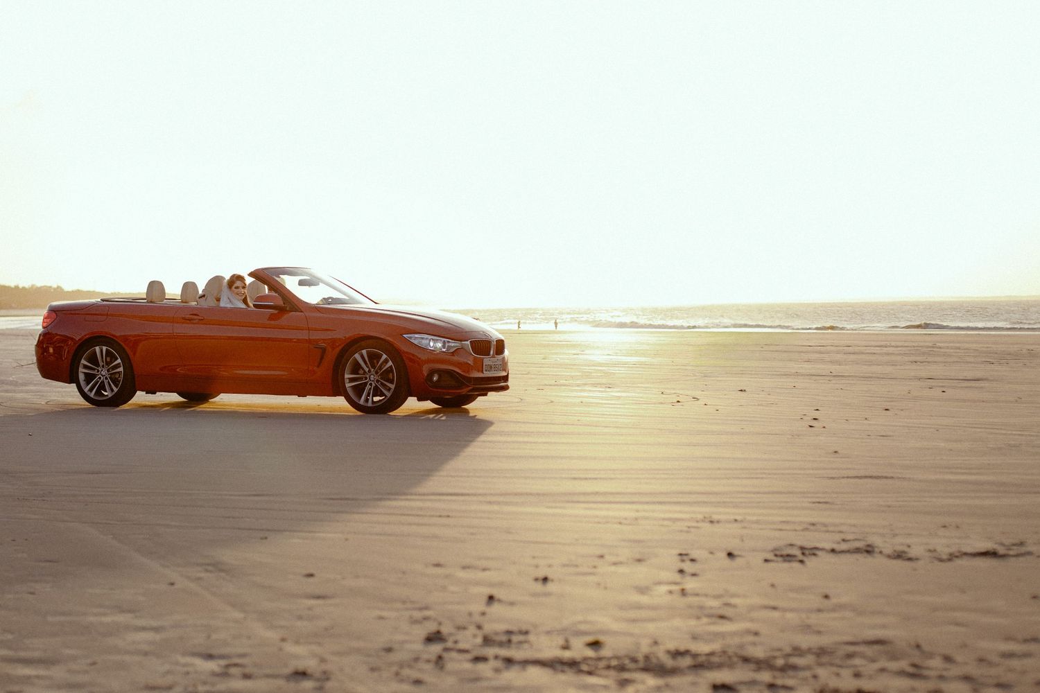 A sporty orange BMW car drifting on a sandy beach at sunset casting a dramatic shadow.