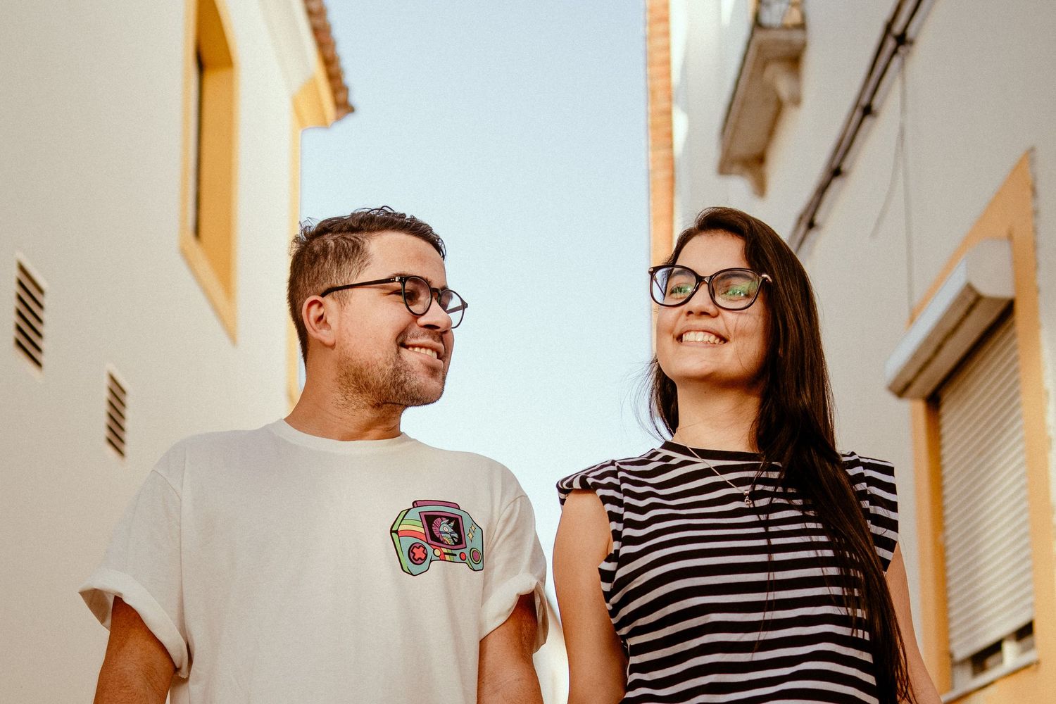 Two people wearing glasses stand together against a white wall, one in a striped shirt and one in a white t-shirt.