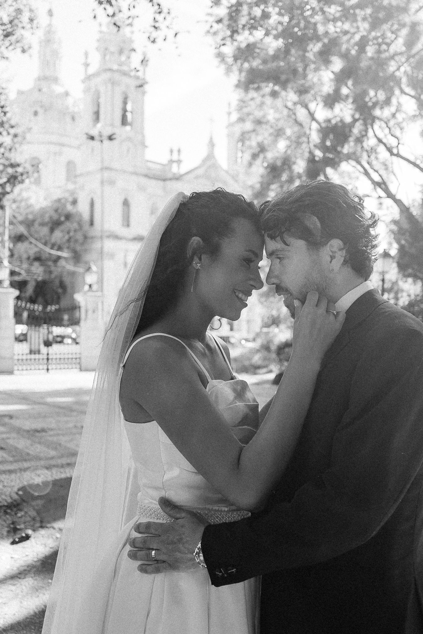 Black and white photo of a bride and groom embracing tenderly outside a church on their wedding day.