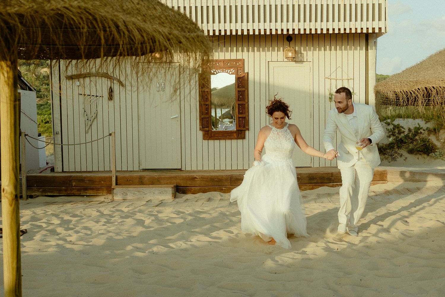 Bride and groom running hand in hand on sandy beach near white wooden building during golden hour.