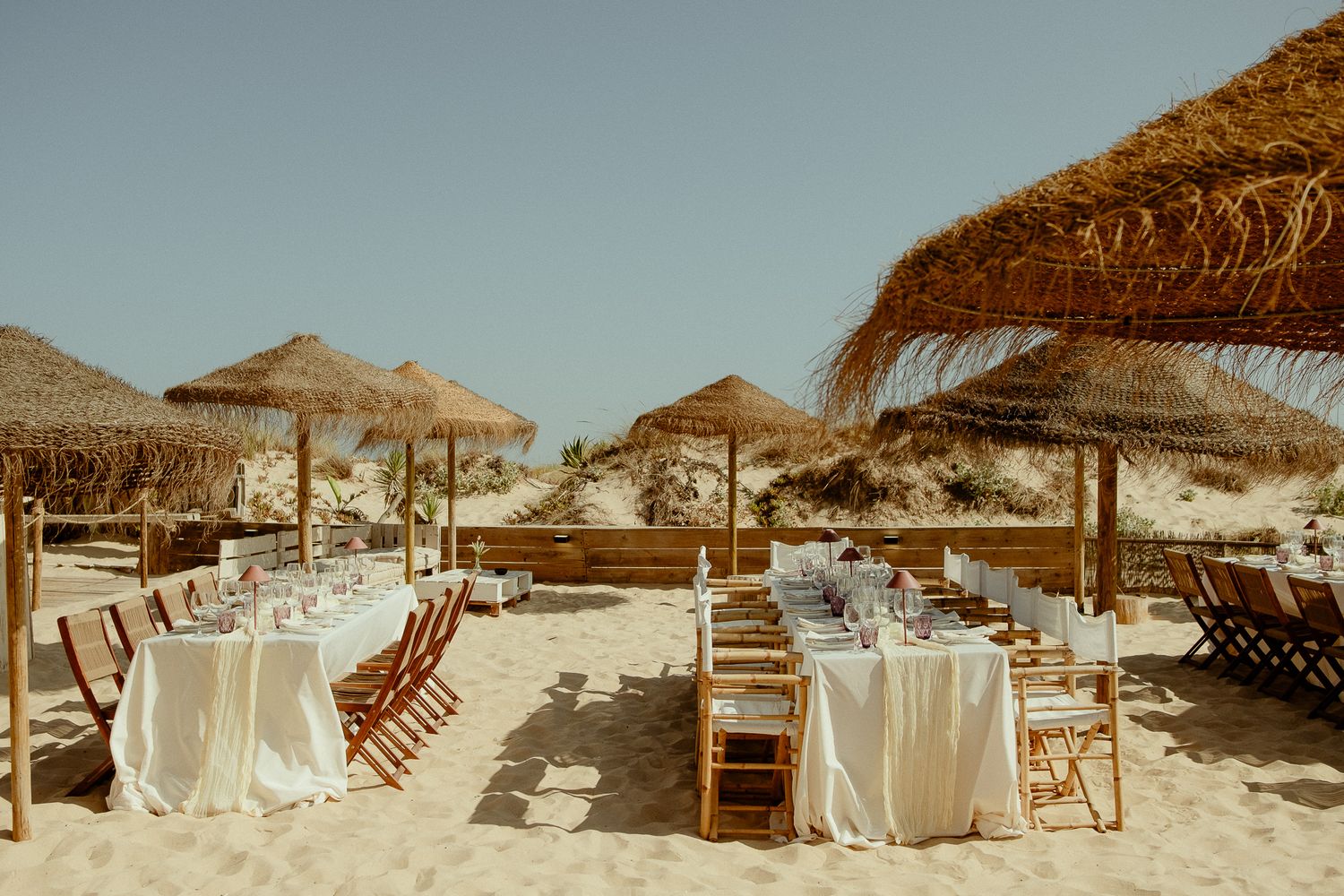 Elegant beach wedding reception setup with white draped tables, wooden chairs, and thatched umbrellas on sandy shore.
