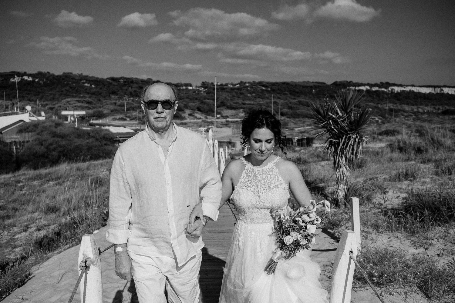 Black and white photo of a bride holding a bouquet walking with a man in sunglasses at an outdoor wedding ceremony.