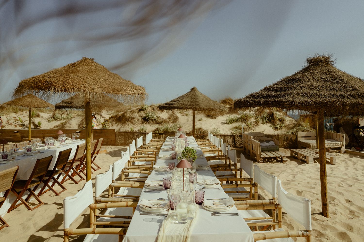 Elegant beach dining setup with long tables, straw umbrellas, and place settings on sandy shore.