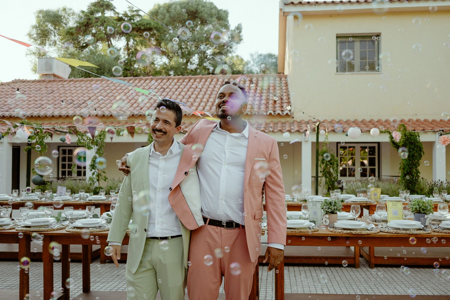 Two men in suits smiling and embracing at an outdoor wedding reception with bubbles floating around them.