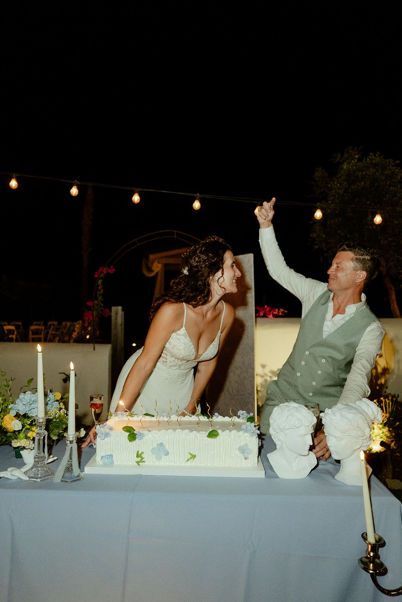 Bride and groom cutting wedding cake outdoors at night, surrounded by candles and string lights.