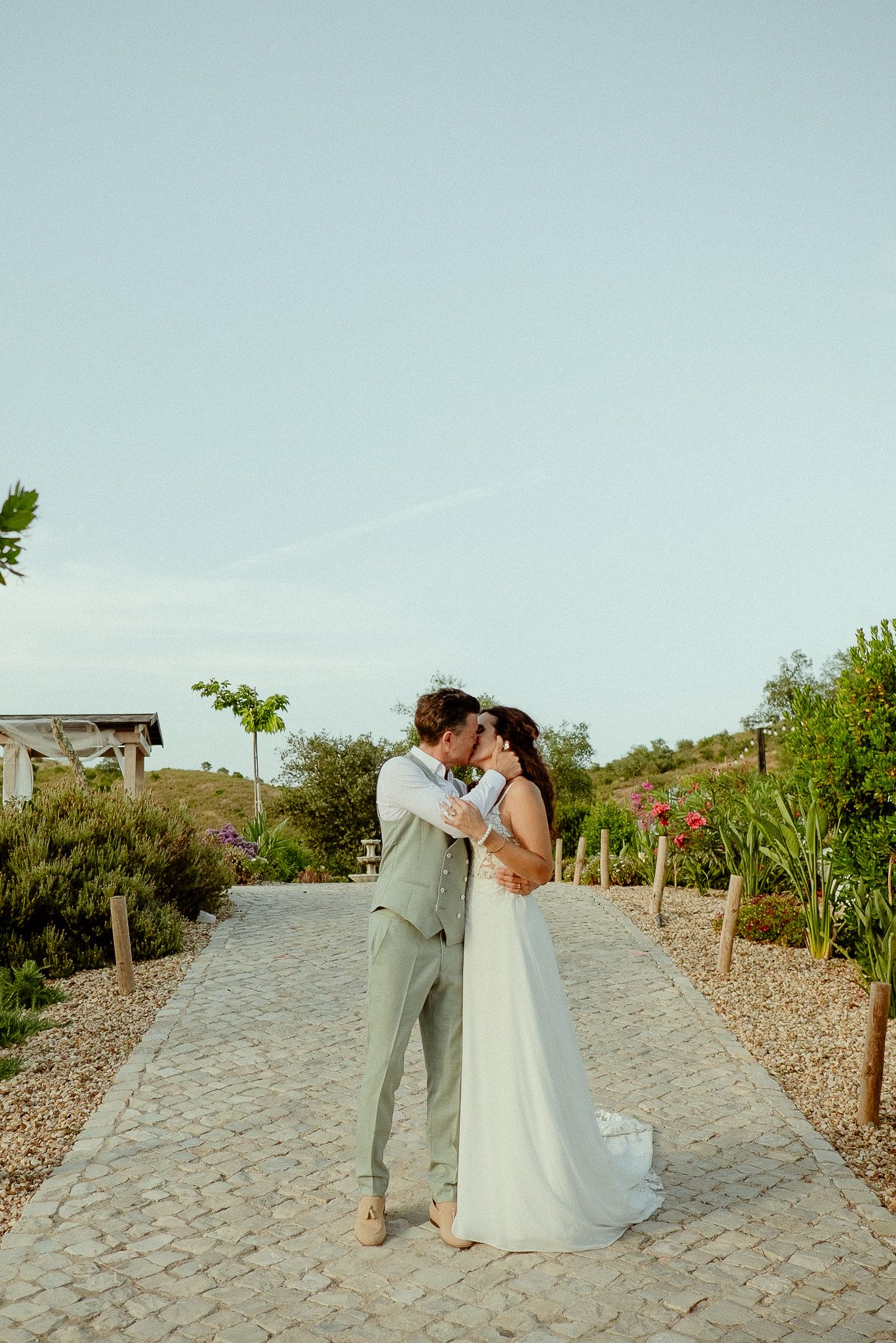 Bride and groom share a romantic kiss on a cobblestone path surrounded by lush gardens on their wedding day.