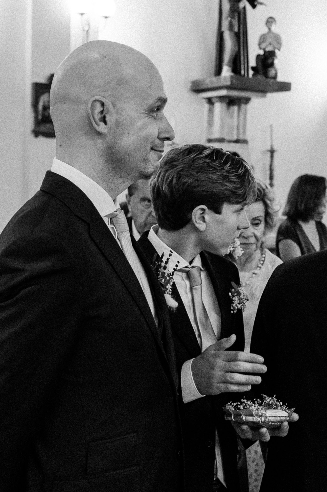 Black and white wedding photo of bald groom and young boy in suits holding a ring pillow in a church.