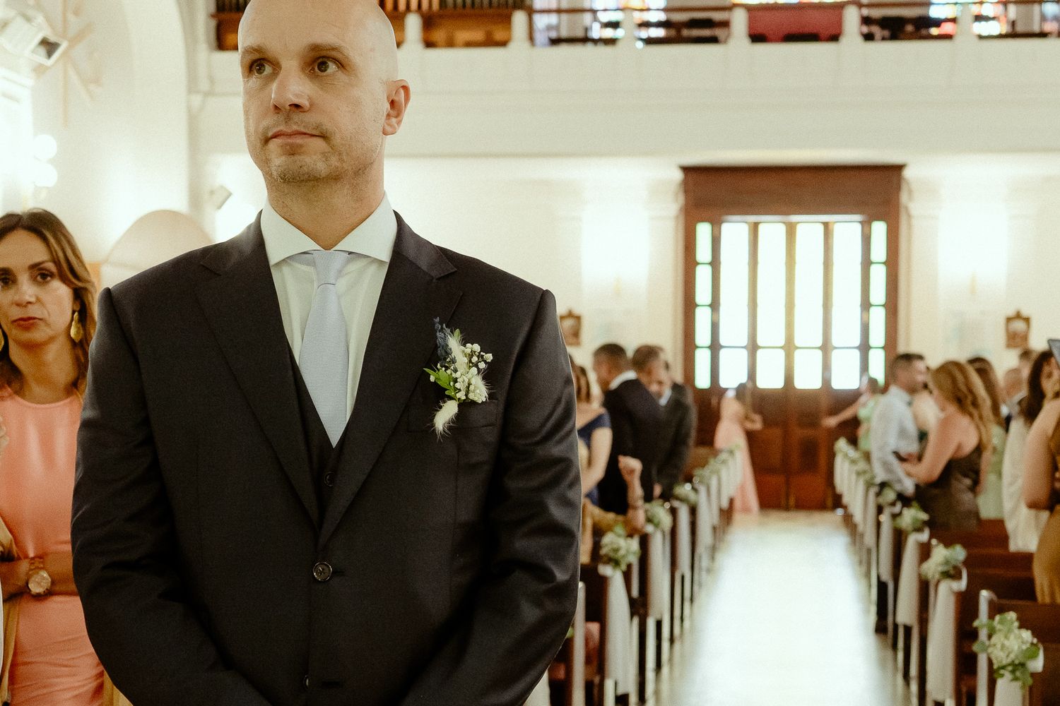 Groom in black suit with boutonniere stands at church altar awaiting bride during wedding ceremony.