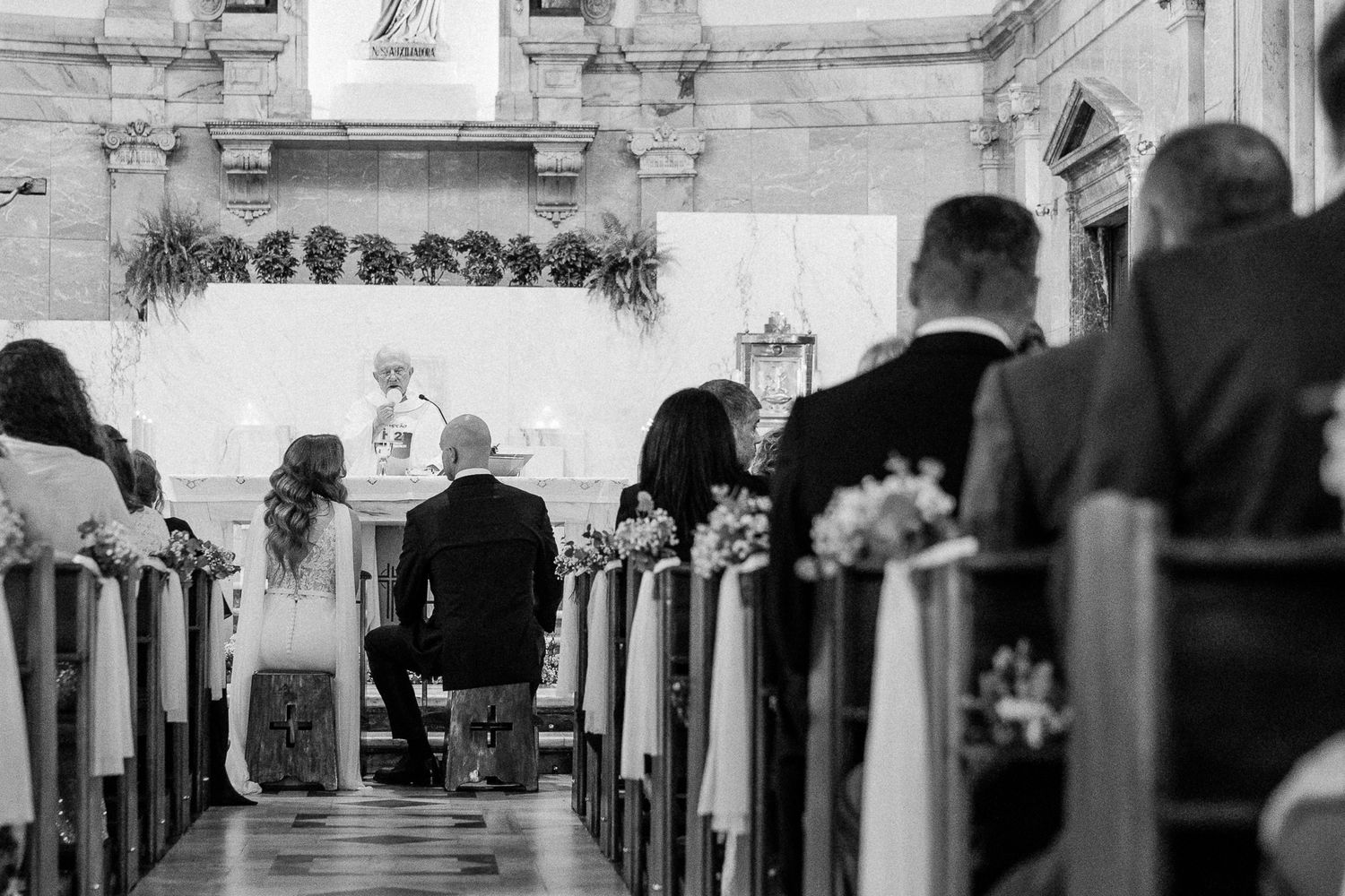 Black and white photo of a wedding ceremony in a church, with guests seated in pews and couple at the altar.