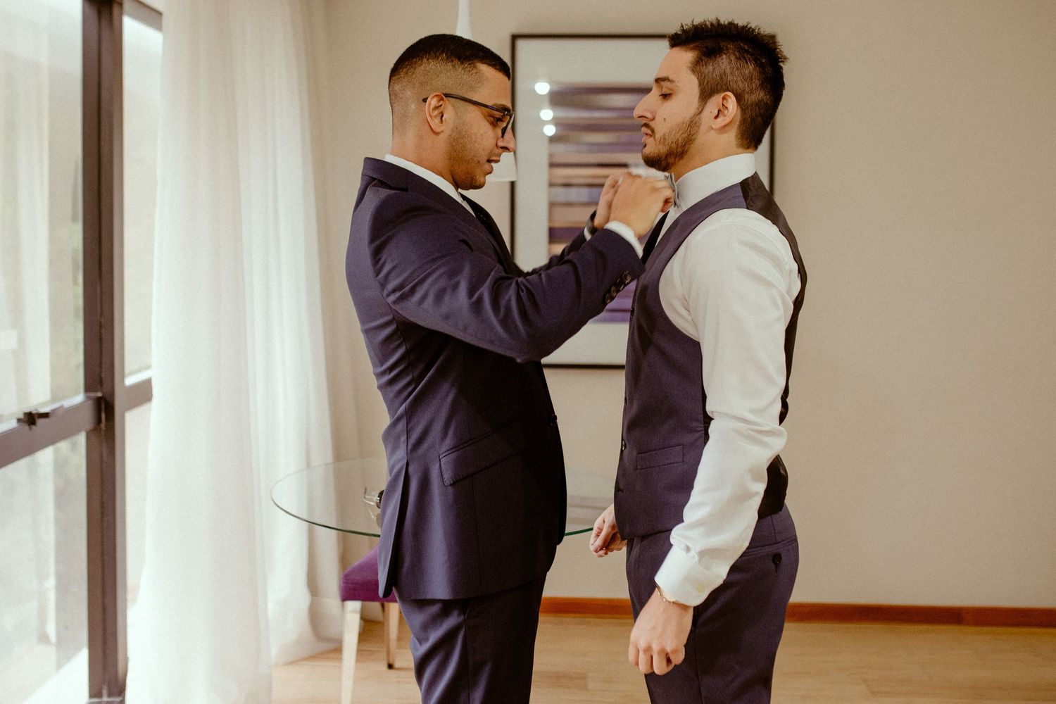 Two people in formal attire help each other get dressed before a special occasion in an indoor setting.